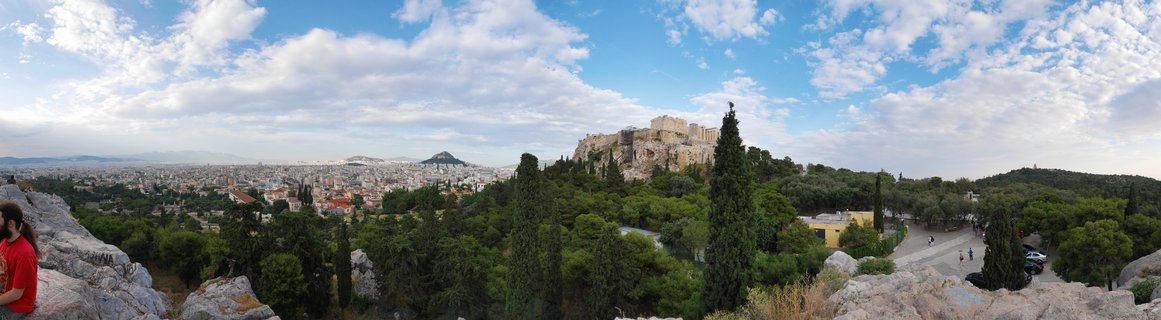Wide panoramic photograph showing the Acropolis of Athens on a rocky hill with the Parthenon visible, surrounded by green trees in the foreground and the sprawling city of Athens extending to distant mountains under a blue sky with white clouds.