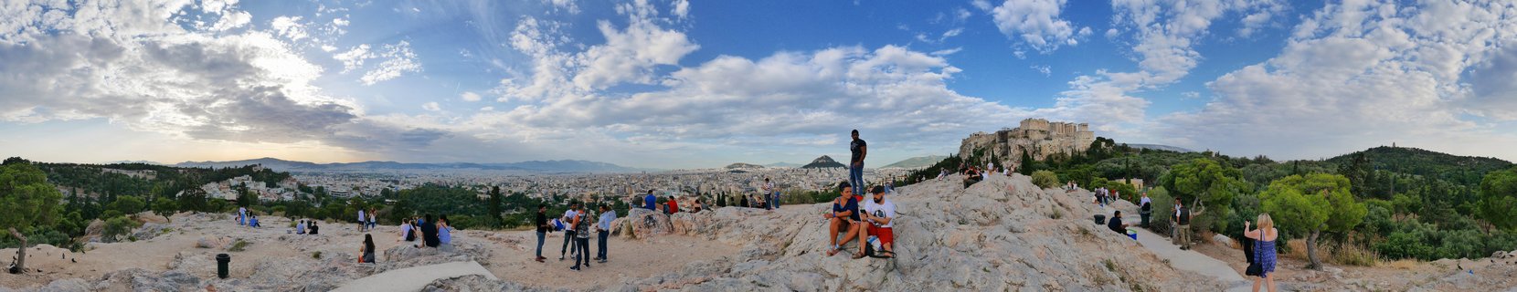 Panoramic view from rocky Areopagus Hill showing tourists scattered across limestone terrain with Athens cityscape and the Acropolis with Parthenon visible in the distance under blue sky with white clouds