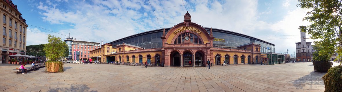 Wide view of Erfurt Central Train Station showing the historic yellow sandstone building with arched entrance, large glass roof structure, and paved plaza with pedestrians under blue sky with scattered clouds