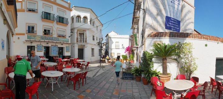 A panoramic view of a narrow cobblestone street in Marbella's old town with white buildings, red café chairs, outdoor restaurant tables, palm trees, and pedestrians under a clear blue sky