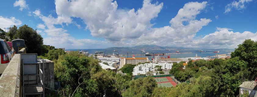 Panoramic view of Gibraltar harbor showing port facilities with cargo ships, cranes, white buildings, tennis courts, and green vegetation under blue sky with white clouds