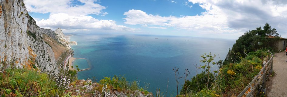Panoramic view of the Rock of Gibraltar limestone cliffs overlooking the Mediterranean Sea with Catalan Bay beach below, ships on the horizon, and vegetation in the foreground under a blue sky with white clouds