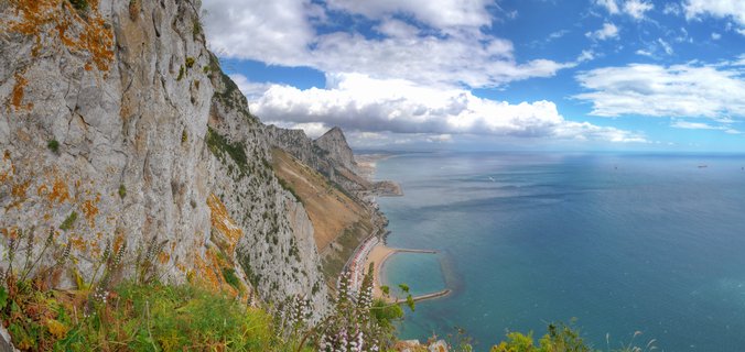 Panoramic view of Gibraltar's limestone cliff face covered in orange lichen, overlooking turquoise Mediterranean waters with a sandy beach and breakwater below under blue sky with white clouds