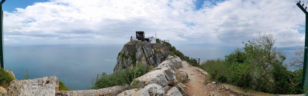 Panoramic landscape showing a rocky mountain summit with a viewing platform building, surrounded by limestone cliffs, Mediterranean vegetation, and expansive blue sea under cloudy sky