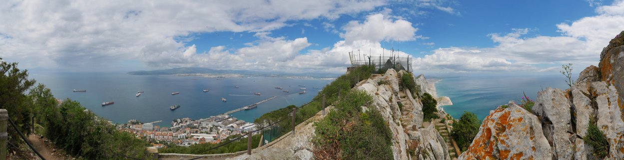 Panoramic landscape view from Rock of Gibraltar showing Mediterranean Sea with cargo ships, harbor, city buildings, limestone cliffs with green vegetation, and blue sky with white clouds