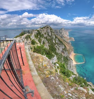 Panoramic landscape showing the Rock of Gibraltar limestone cliffs with green vegetation, a red viewing platform with black railing in foreground, turquoise Mediterranean Sea, sandy beach with breakwater, and blue sky with white clouds