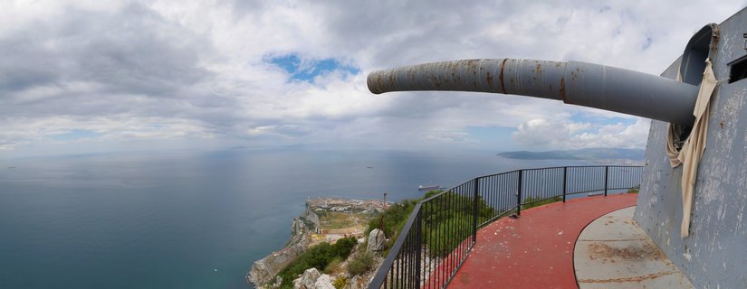 A large grey naval cannon extends from a concrete battery emplacement on the right, overlooking the Mediterranean Sea from Gibraltar's clifftop with a red walkway and black railing in the foreground