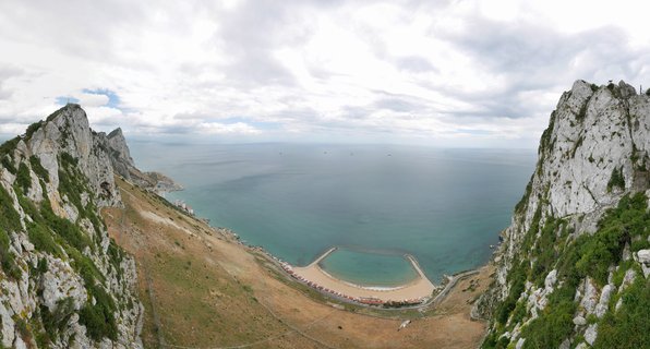 Aerial view showing limestone cliffs of Rock of Gibraltar on both sides framing Catalan Bay beach with rectangular swimming pool and Mediterranean Sea under cloudy sky