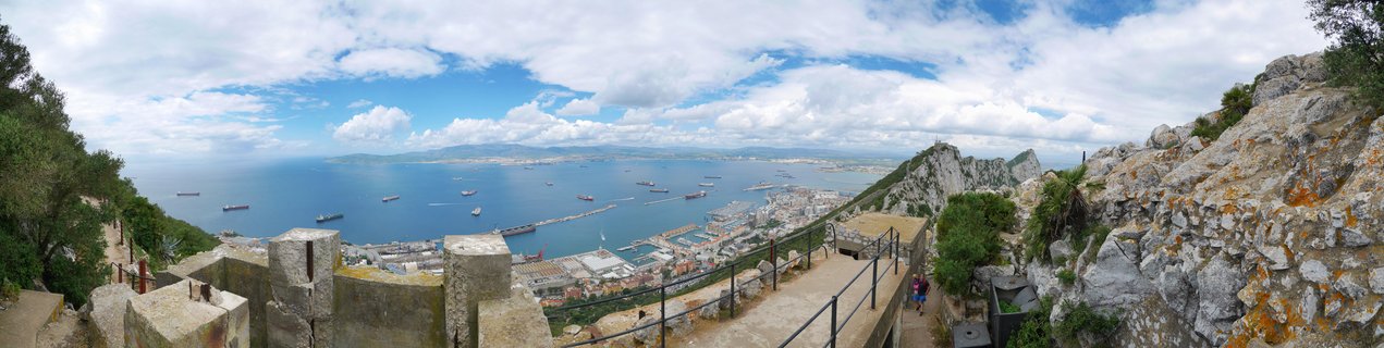 Panoramic landscape showing Gibraltar harbor from elevated stone fortifications with the Rock formation, cargo ships in blue bay waters, and distant Spanish coastline under cloudy sky