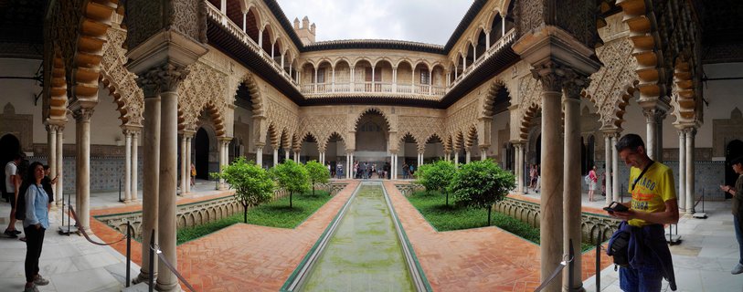 Historic palace courtyard with central water feature, surrounded by two-story arcades with ornate horseshoe arches, marble columns, carved stucco walls, and small orange trees in garden beds