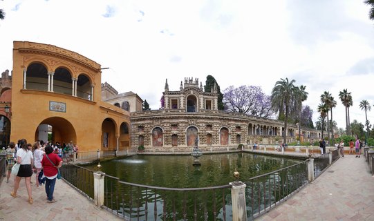 Historic palace complex in Seville with ornamental pool, fountain, arched galleries, and tourists walking on stone pathways surrounded by palm trees