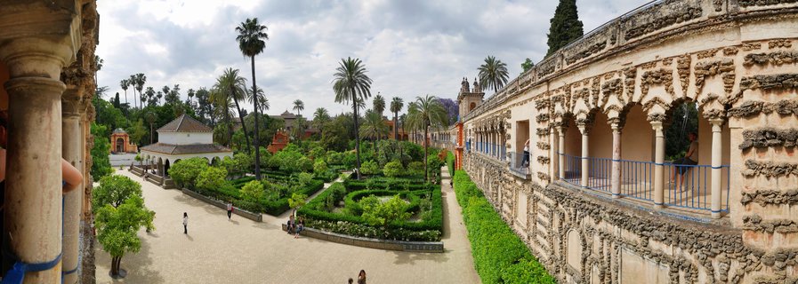 Elevated view of the Alcázar palace gardens in Seville showing ornate stone arches along the right, formal geometric hedges, palm trees, and visitors walking on gravel pathways under cloudy skies