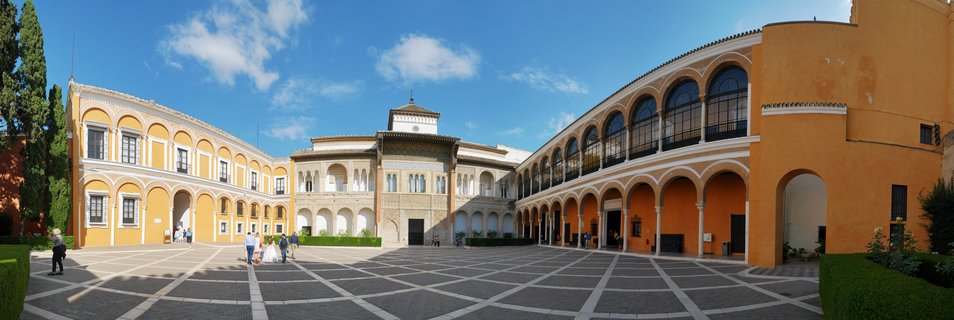 Wide courtyard of Real Alcázar Palace showing orange buildings with white arched colonnades, geometric stone paving, visitors walking, cypress trees, and blue sky with scattered clouds