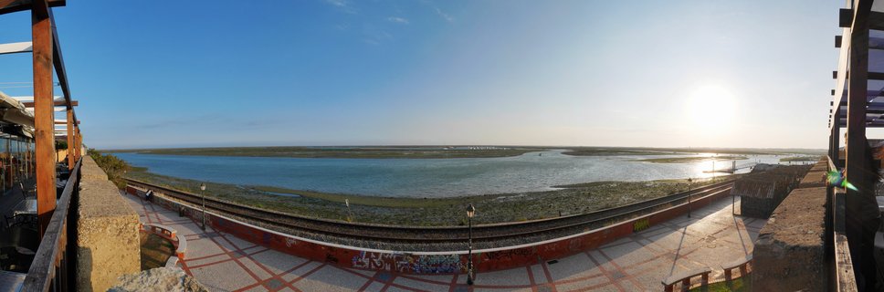Panoramic coastal landscape showing a lagoon with railway tracks, tiled walkway, and wooden terrace structures under a clear blue sky at sunset in Faro, Portugal