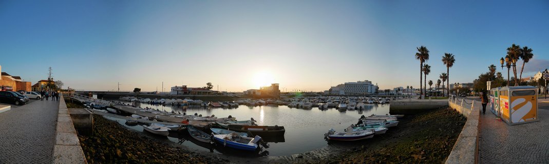 Panoramic view of a marina harbor at sunset with numerous small boats docked in calm water, palm trees on the right, cobblestone walkways on both sides, and buildings in the background under a clear sky with the sun low on the horizon