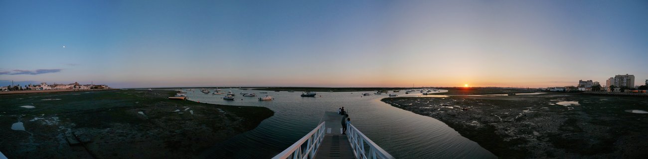 Panoramic sunset view over coastal marsh estuary with wooden pier, anchored boats, and distant buildings along the horizon in Faro, Portugal