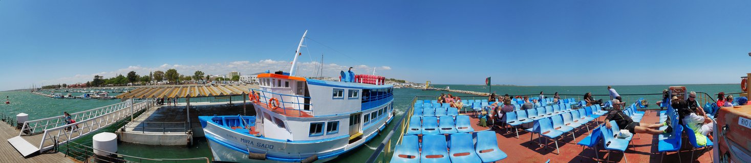 A white and blue ferry boat named Mira Sado docked at a wooden pier with passengers seated on blue chairs on the open deck under a clear blue sky