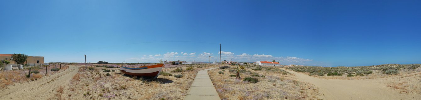 A small white and orange fishing boat sits on dry grassy coastal ground with dirt paths, concrete walkway, distant white buildings with red roofs, and clear blue sky in Culatra, Portugal