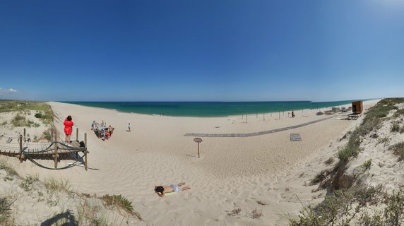 Wide beach scene showing white sand dunes, turquoise ocean, wooden walkways, and people sunbathing and walking on a sunny day in Portugal