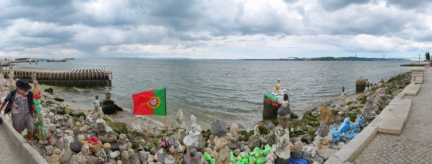 Panoramic waterfront scene showing painted stone sculptures and cairns along rocky Tagus River shoreline in Lisbon, Portugal, with Portuguese flag and 25 de Abril Bridge visible in distance under cloudy sky