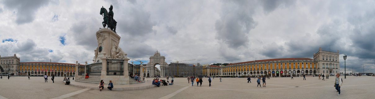 Wide panoramic view of Praça do Comércio showing the equestrian statue of King José I on a stone pedestal, the Rua Augusta triumphal arch in the background, yellow historic buildings with arcades, and people walking across the stone plaza under cloudy skies