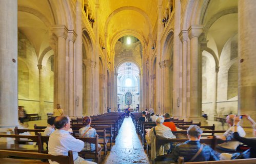 Interior view of a cathedral nave with stone columns, rounded arches, wooden pews with seated visitors, and a central aisle leading to an altar in the distance