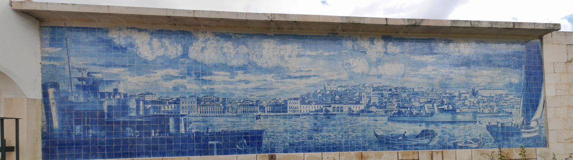 Large blue and white Portuguese azulejo tile mural showing Lisbon harbor with ships, boats, waterfront buildings, and cloudy sky mounted on outdoor wall