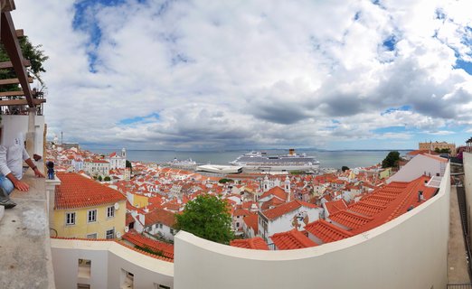 Panoramic cityscape of Lisbon showing red-tiled rooftops of historic buildings, white curved walls in foreground, cruise ship docked at port, Tagus River, and cloudy sky with blue patches