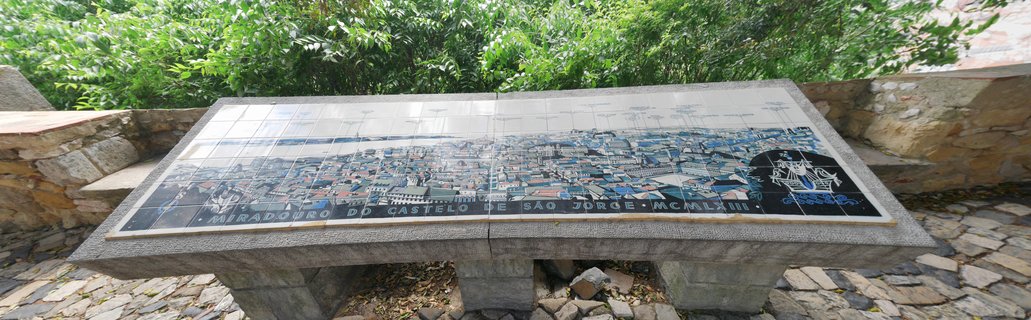 Stone-mounted azulejo tile panel showing illustrated Lisbon cityscape at São Jorge Castle viewpoint with green foliage above and cobblestone ground below