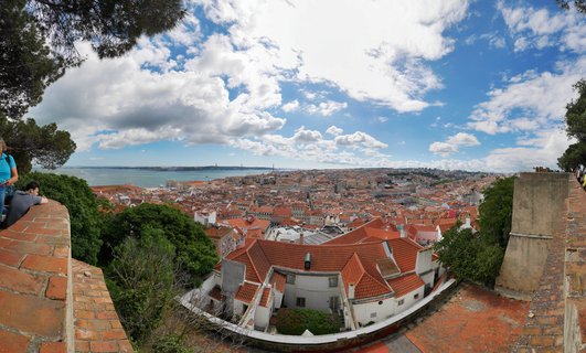 Elevated panoramic cityscape photograph of Lisbon Portugal featuring dense urban fabric of white stucco buildings with terracotta clay tile roofs, foreground stone brick viewing terrace with tourists, mature pine trees framing composition, Tagus River estuary visible at horizon, 25 de Abril suspensi
