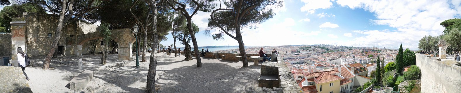 Panoramic view from castle grounds showing Lisbon cityscape with red-tiled roofs, stone archways, pine trees, and the Tagus River in the distance under blue sky with clouds