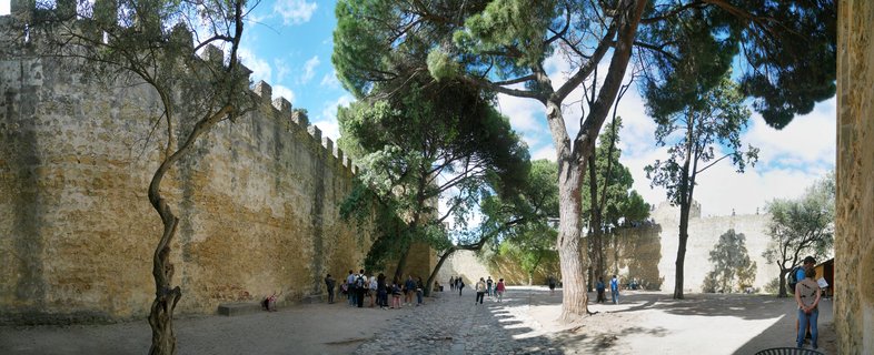 Medieval stone castle walls with crenellated battlements, pine trees, and tourists walking on cobblestone pathway under blue sky with white clouds