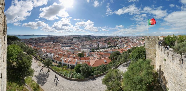 Panoramic cityscape of Lisbon showing red-tiled rooftops, stone castle walls with Portuguese flag, cobblestone pathways with visitors, green trees, Tagus River and bridge in the background under blue sky with white clouds