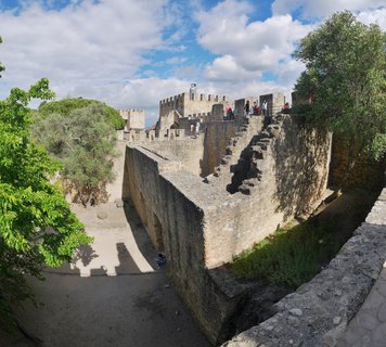 Medieval stone castle fortress with crenellated walls and towers, tourists walking on ramparts, green trees, blue sky with white clouds, Lisbon Portugal