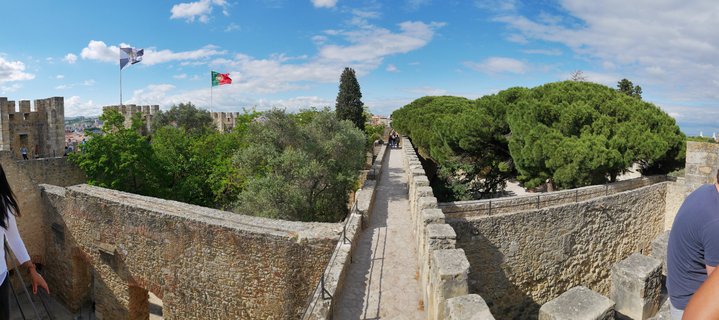 Stone castle battlements with a walkway running along medieval fortification walls, Portuguese flag visible, green trees, blue sky with white clouds