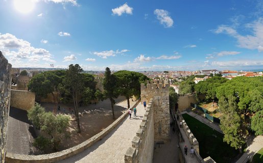 Aerial view of medieval castle stone walls with crenellations, tourists walking on battlements, green pine trees in courtyard, Lisbon cityscape with red rooftops in background under blue sky with white clouds