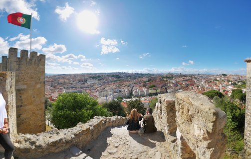 Two people sitting on stone castle walls overlooking Lisbon cityscape with Portuguese flag flying from tower under blue sunny sky