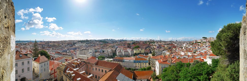 Wide panoramic cityscape of Lisbon showing dense urban buildings with red-tiled roofs under blue sky with white clouds, viewed from elevated castle viewpoint