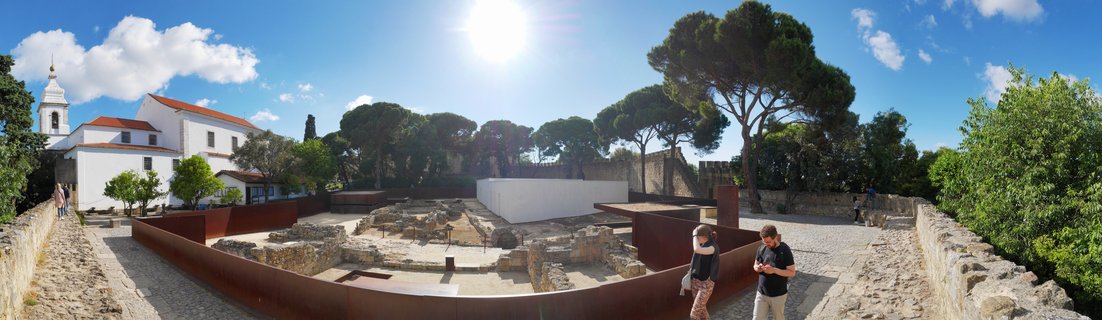 Panoramic view of archaeological ruins with stone foundations enclosed by rust-colored metal barriers at São Jorge Castle, with white church building, medieval stone walls, pine trees, and tourists visiting the historic site in Lisbon under blue sky