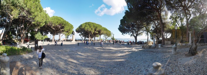 Panoramic view of archaeological garden at São Jorge Castle in Lisbon showing ancient stone column ruins, Mediterranean umbrella pine trees, traditional Portuguese cobblestone pavement, and distant Tagus River under blue sky with scattered clouds during golden hour