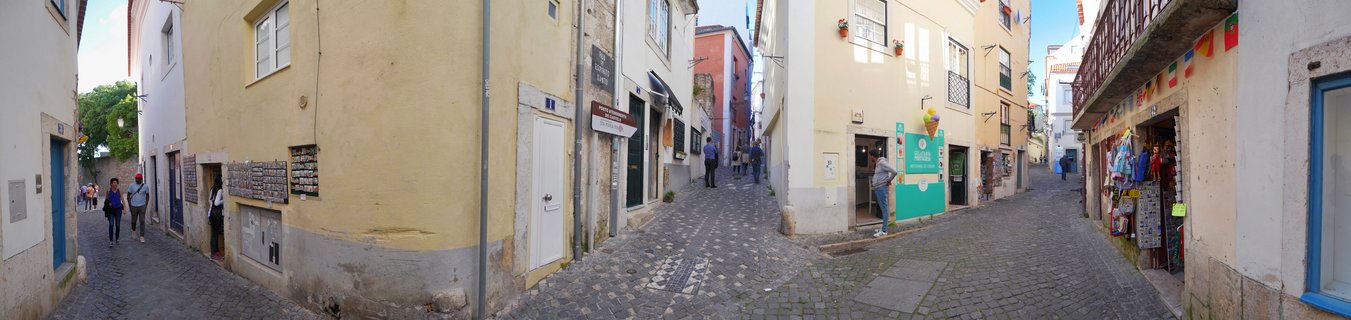 A narrow cobblestone street in Lisbon with yellow and white buildings on both sides, people walking, shop fronts with merchandise, and Portuguese flags visible