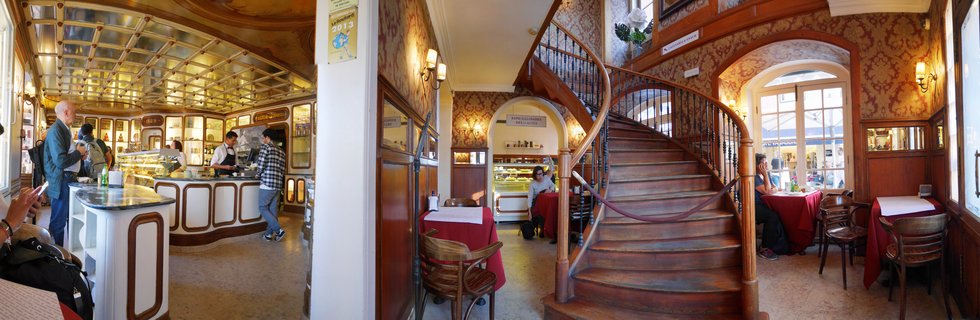 Interior of historic Portuguese café with wooden spiral staircase, ornate wallpaper, pastry display cases, customers at tables with red tablecloths, and Belle Époque architectural details