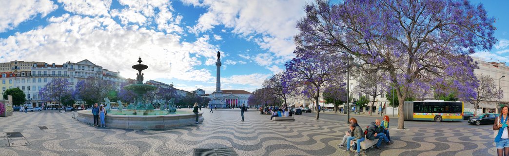 Panoramic view of Rossio Square in Lisbon showing ornate fountain, tall column monument, purple flowering jacaranda trees, Portuguese wave-pattern pavement, and people in a historic urban plaza under partly cloudy sky