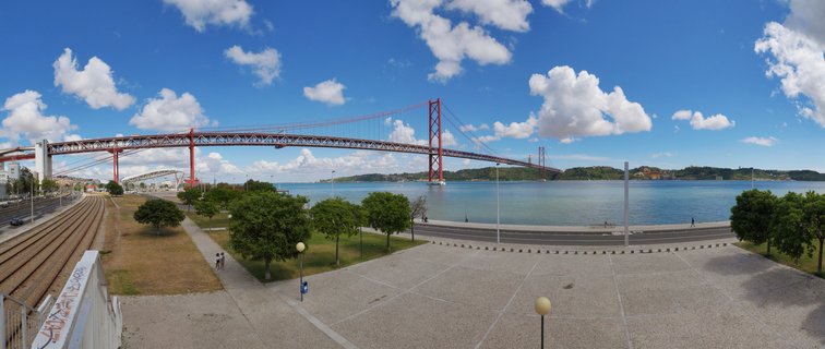 Red suspension bridge spanning wide blue river with paved waterfront plaza, trees, and train tracks visible in foreground under partly cloudy sky
