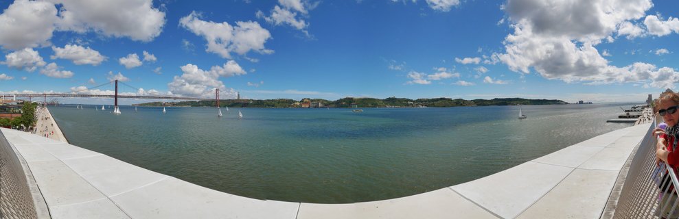 Panoramic view of the red 25 de Abril Bridge crossing the Tagus River in Lisbon with sailboats on the water and a stone waterfront promenade in the foreground under a blue sky with white clouds