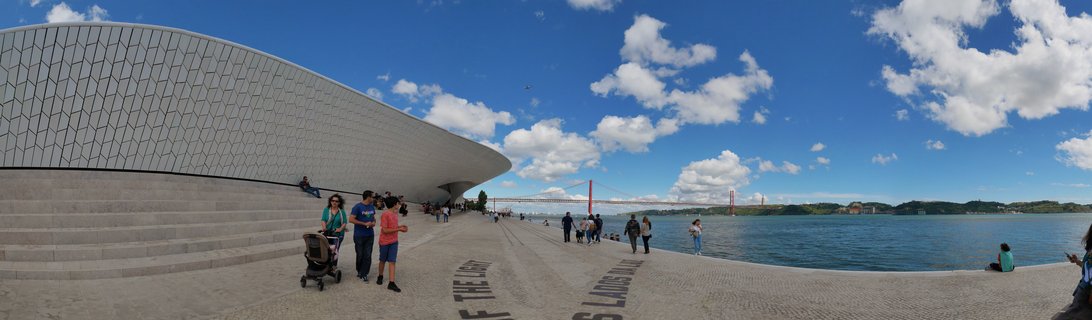Wide panoramic view of Lisbon waterfront showing the white curved MAAT museum building with geometric tile pattern on the left, people walking on stone promenade, and the red 25 de Abril Bridge spanning the Tagus River under blue sky with white clouds