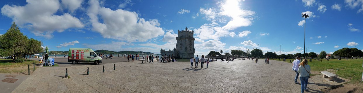 Wide view of Belém Tower in Lisbon with tourists walking on stone plaza under blue sky with white clouds, food truck and trees visible on the sides