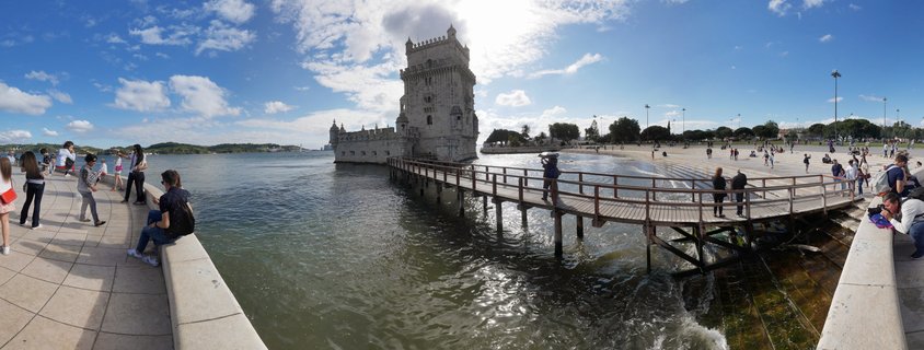 Panoramic view of Belém Tower on the Tagus River in Lisbon with tourists on a wooden walkway and riverside promenade under a blue sky with clouds