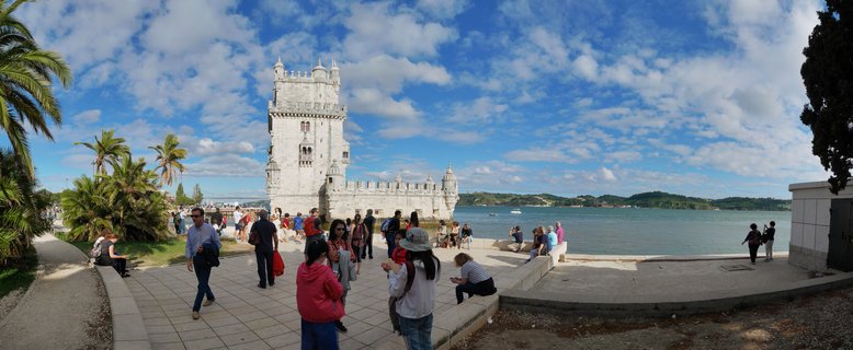 The Belém Tower, a historic stone fortress with ornate architecture, stands on the waterfront in Lisbon with tourists walking on a paved promenade and the Tagus River in the background under a blue sky with scattered clouds