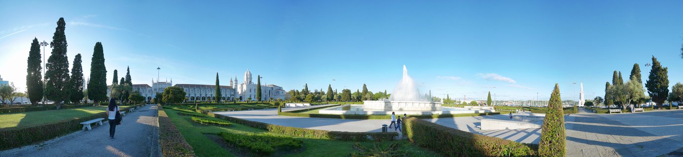 Wide panoramic view of a large plaza with fountain, manicured gardens, and historic white monastery building under clear blue sky in Lisbon